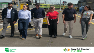 A group of six diverse young professionals walking and chatting on a sunny train station platform. A green train is visible in the background, and the image includes logos for TFI and Irish Rail at the bottom.